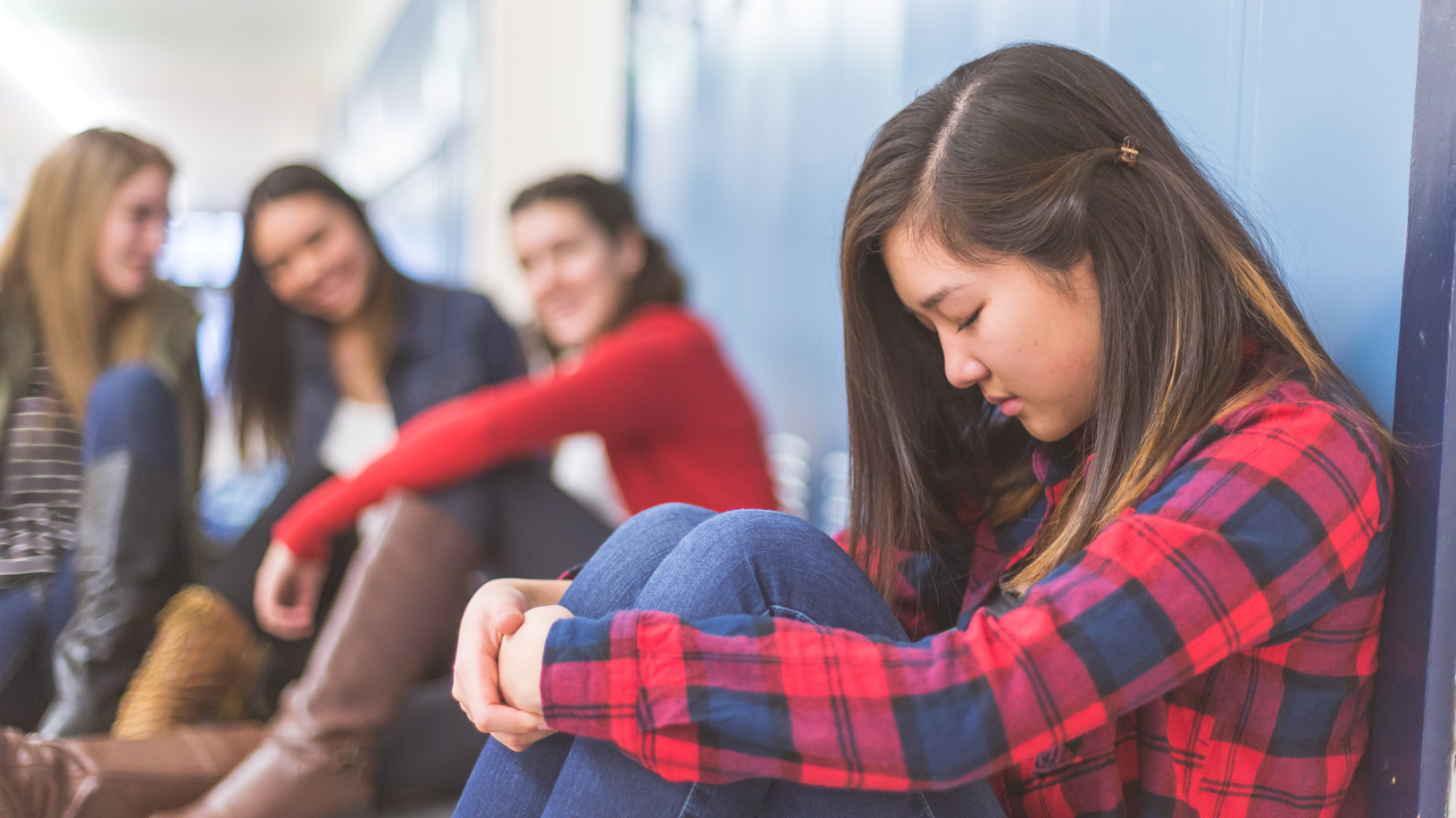 Three girls in the background talking amongst themselves and laughing at girl sitting by herself against the locker.