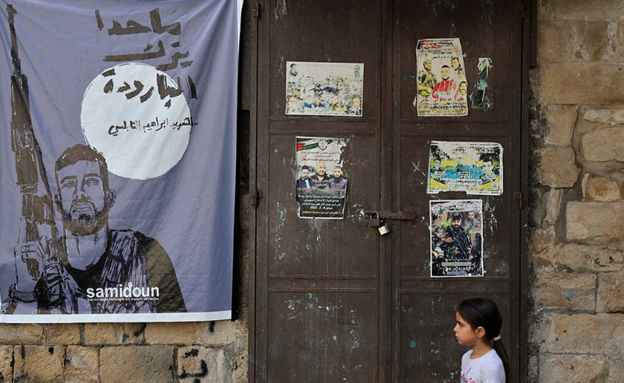 A Palestinian girl stands near a poster of Ibrahim al-Nabulsi, a commander of the Al-Aqsa Martyrs' Brigade, one of the main terrorist groups present in the West Bank operating under the ruling Fatah party, who was killed by Israeli troops in August 2022. The poster bears the Samidoun logo