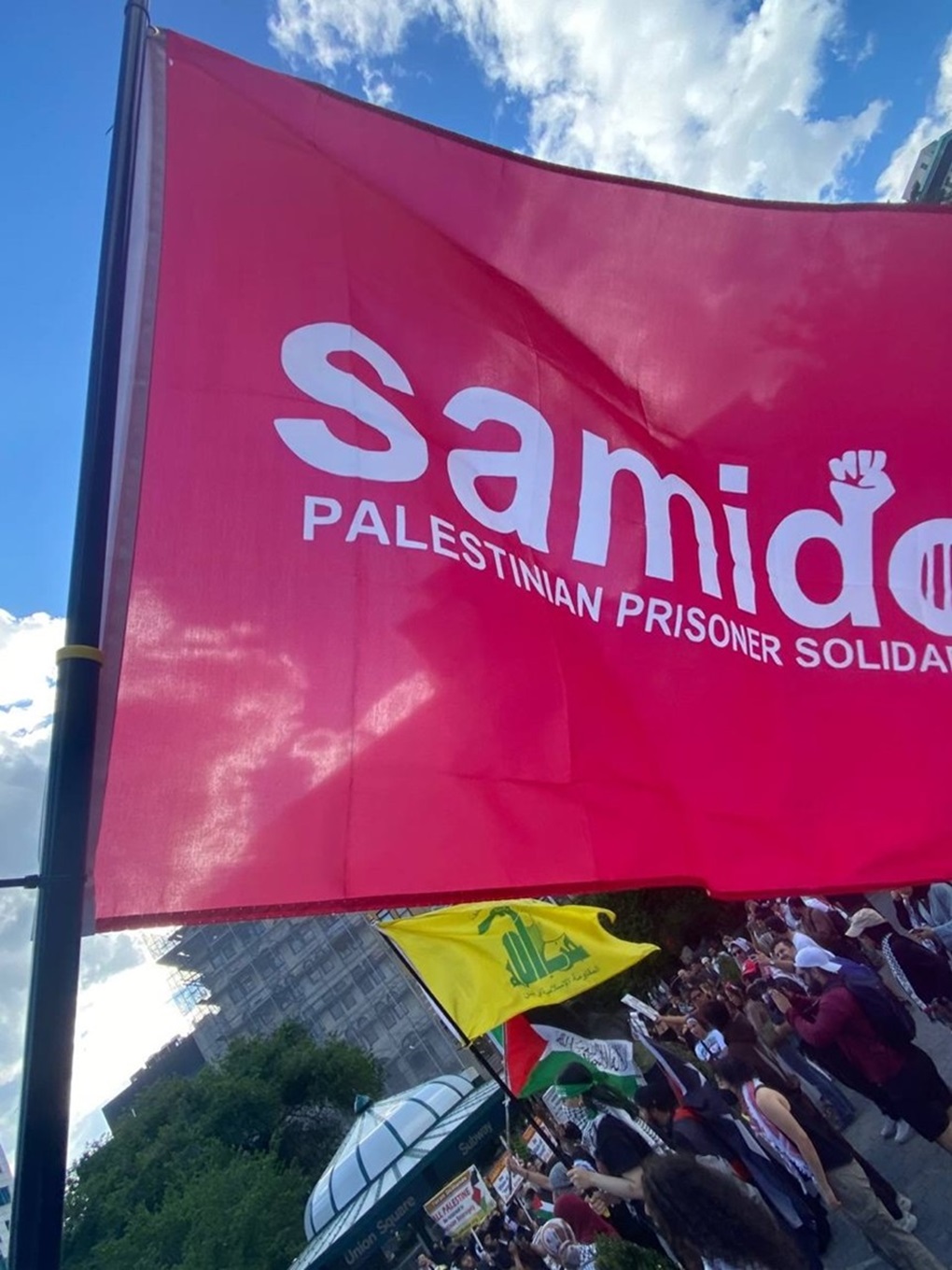 A Samidoun flag is held at a June 10, 2024 anti-Israel rally in New York City. A Hezbollah flag can be seen in the background.