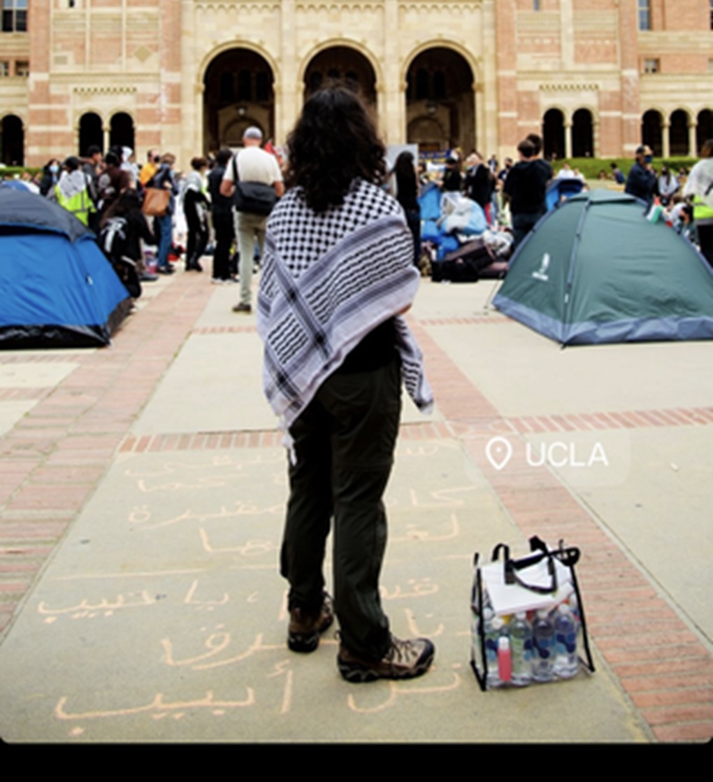 A protestor at the UCLA encampment stands over chalking that reads, “Oh Qassam [Hamas's military wing], burn Tel Aviv